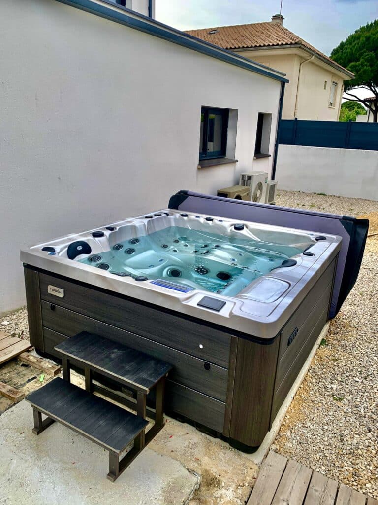 Grand spa extérieur Vendôm moderne avec coque claire et habillage gris foncé, sur socle en béton et gravier, à côté d'une façade blanche.