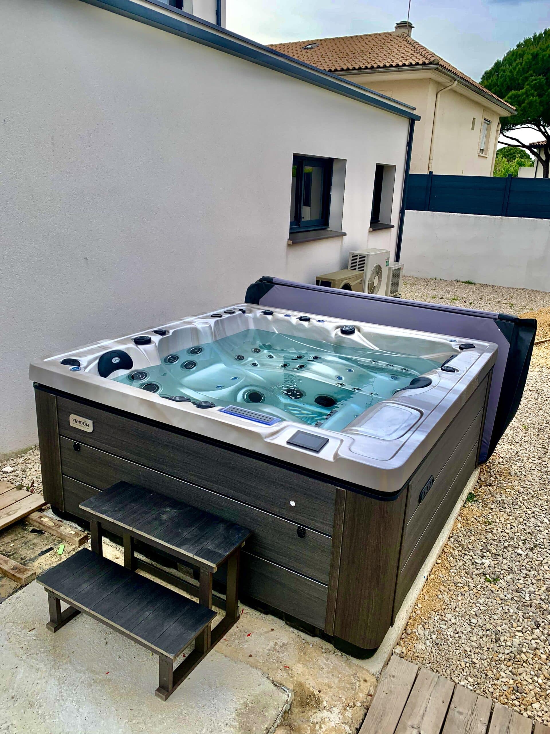 Grand spa extérieur Vendôm moderne avec coque claire et habillage gris foncé, sur socle en béton et gravier, à côté d'une façade blanche.