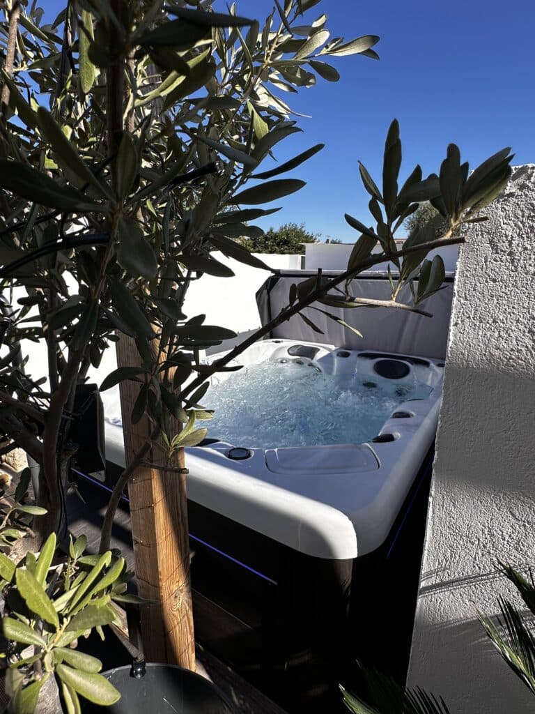 Jacuzzi extérieur sous un olivier, moment de pure détente Jacuzzi moderne en extérieur, l'eau bouillonne sous un ciel bleu éclatant. Encadré par un olivier et un mur blanc texturé.