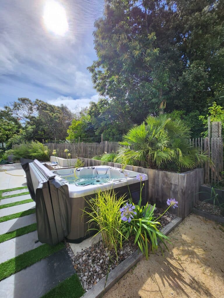 Spa extérieur moderne: Aménagement jardin et terrasse bois Jacuzzi extérieur sous le soleil, intégré à un aménagement paysager. Jardinières en bois, palmiers et Agapanthes violettes bordent la terrasse.