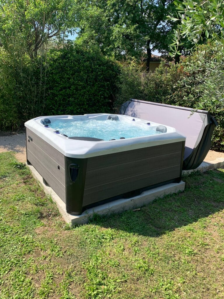Spa de jardin en marche : moment de détente assuré. Bain à remous gris et blanc, jets activés, installé sur l'herbe au milieu de haies denses et d'arbres feuillus. La couverture est relevée.