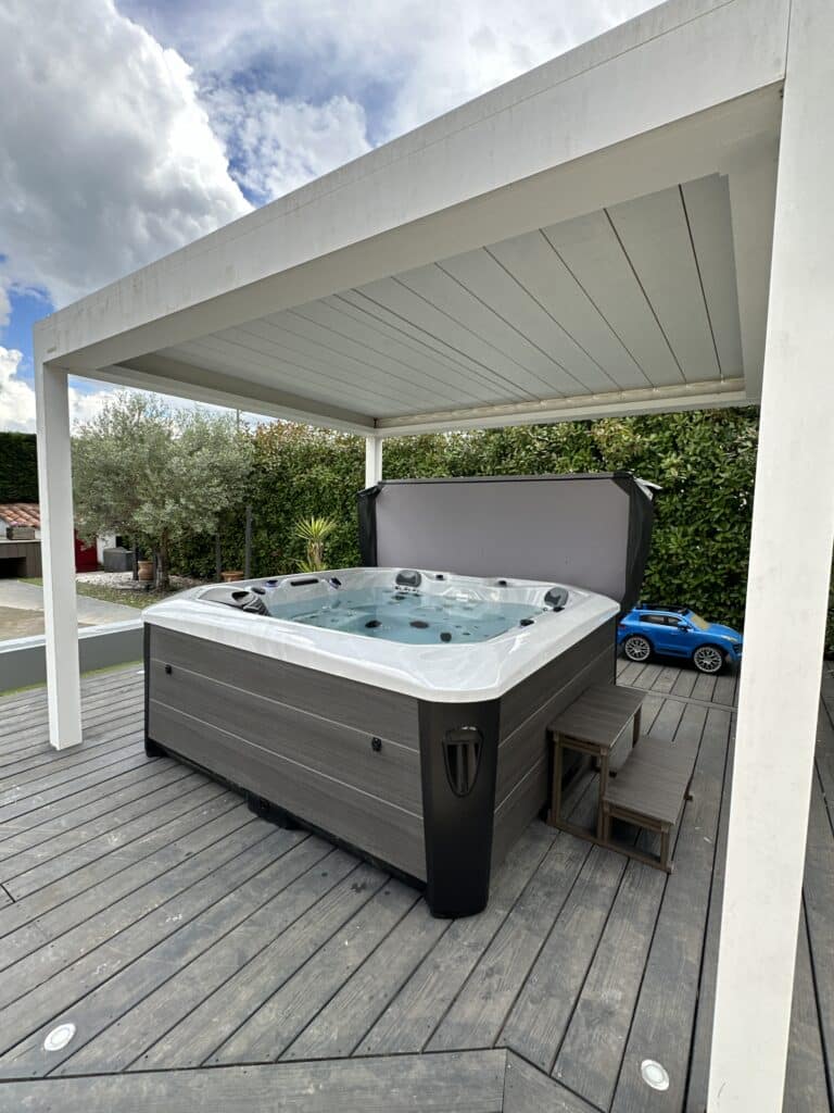 Spa de jardin sous pergola : Aménagement extérieur parfait. Spa de jardin moderne gris sous pergola blanche. Aménagement extérieur luxueux sur terrasse bois pour la détente, ciel nuageux.