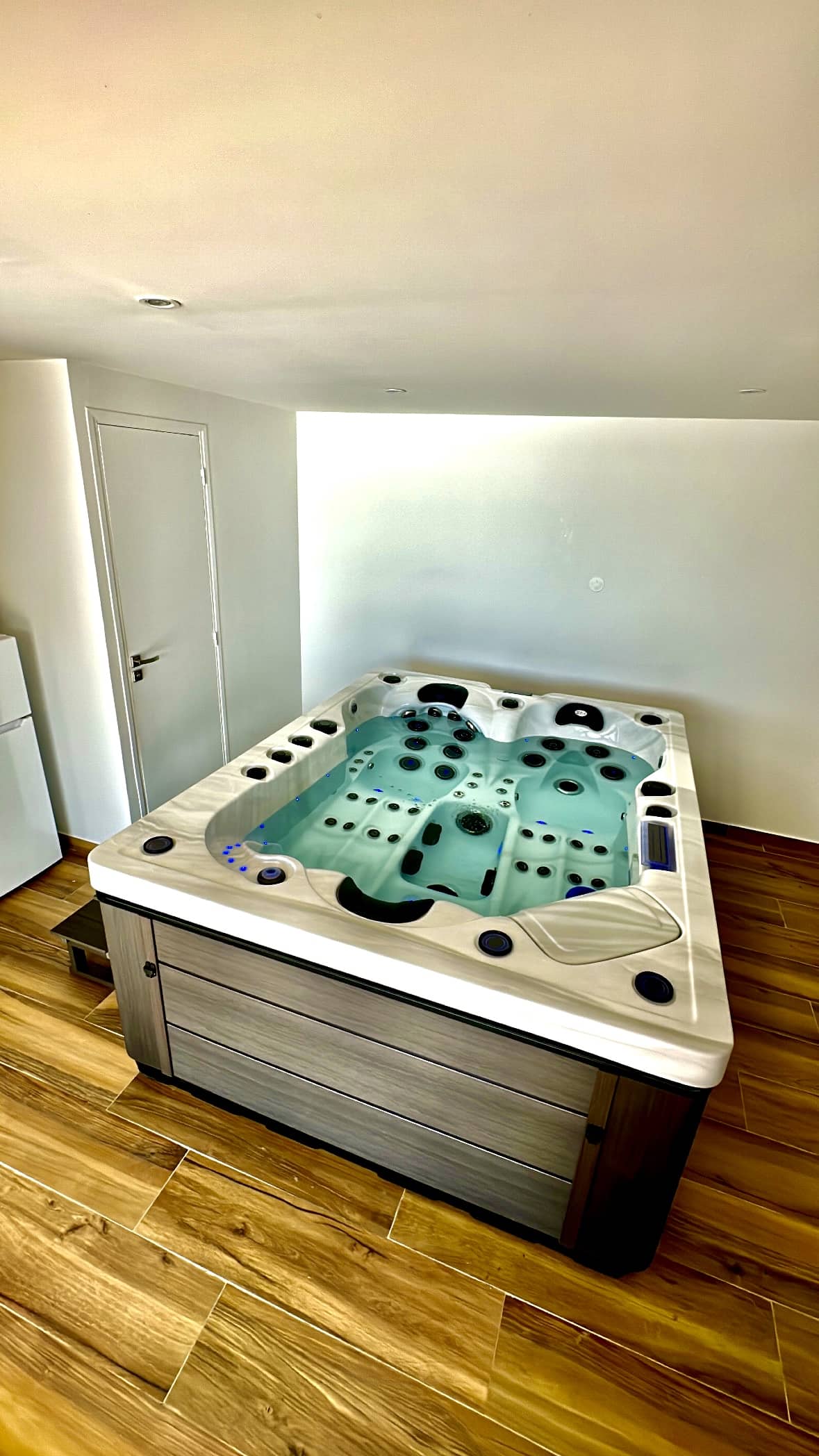 Grand spa à remous intérieur moderne, éclairé par LED bleues, sur sol en carrelage imitation bois. Ambiance lumineuse et épurée.