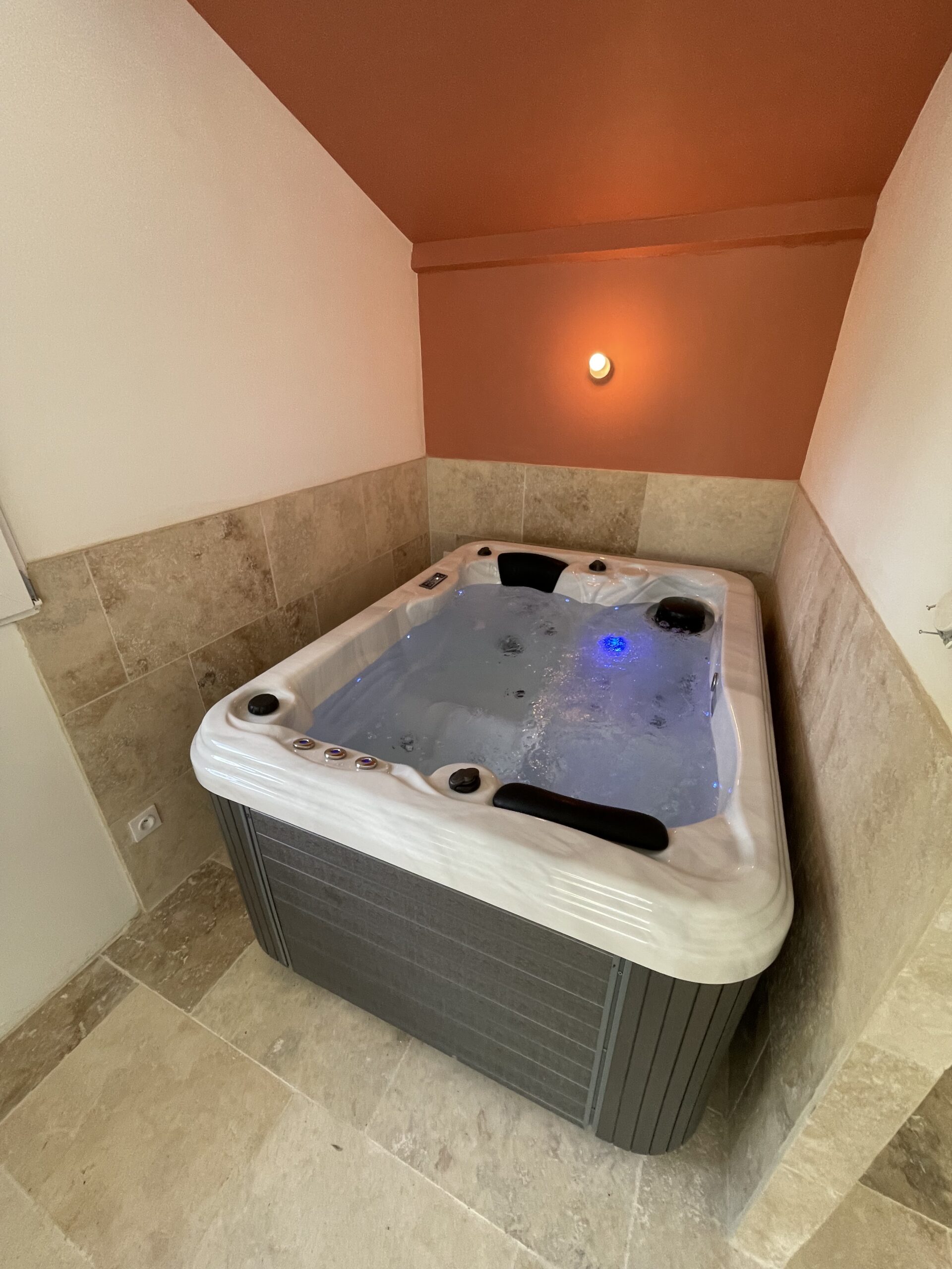 Spa d'intérieur moderne avec coque blanche et revêtement gris. Carrelage en pierre beige et mur d'accent terracotta éclairé, ambiance chaleureuse.