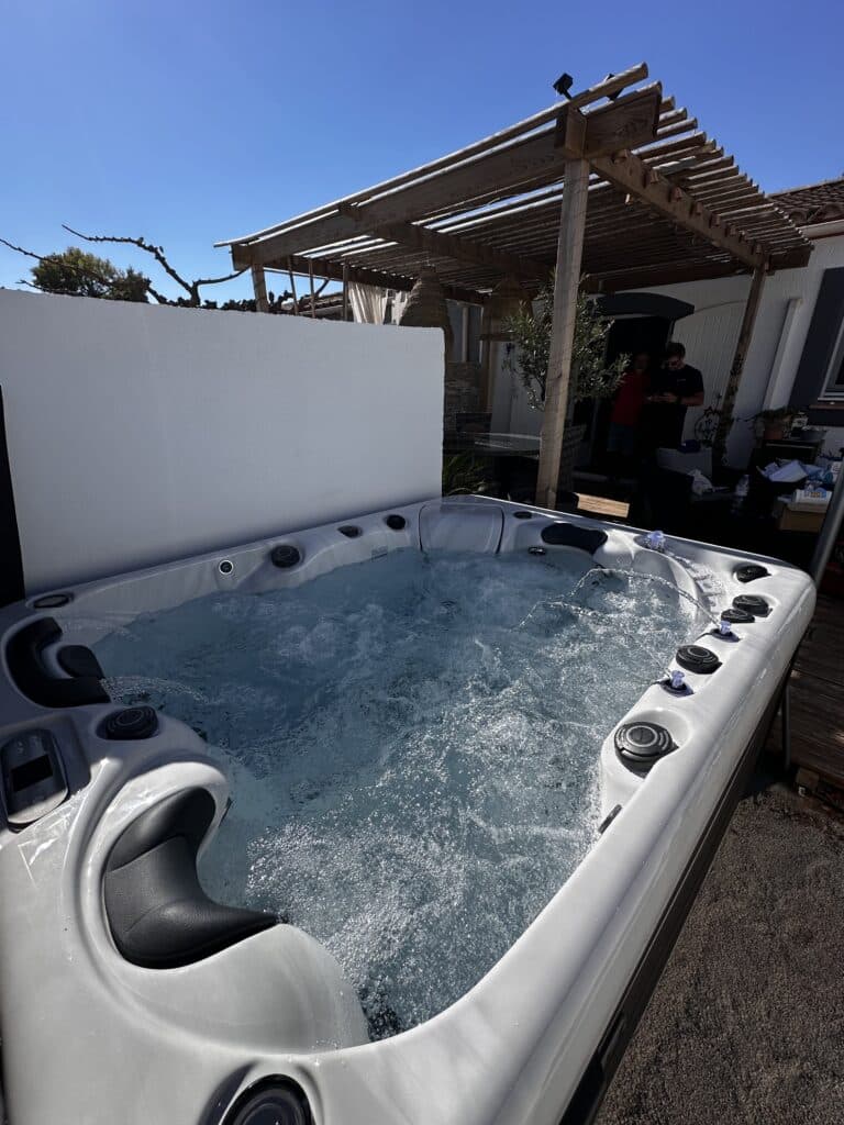 Spa jacuzzi extérieur blanc bouillonnant sur une terrasse. Ciel bleu éclatant, pergola en bois et mur de séparation ensoleillés.