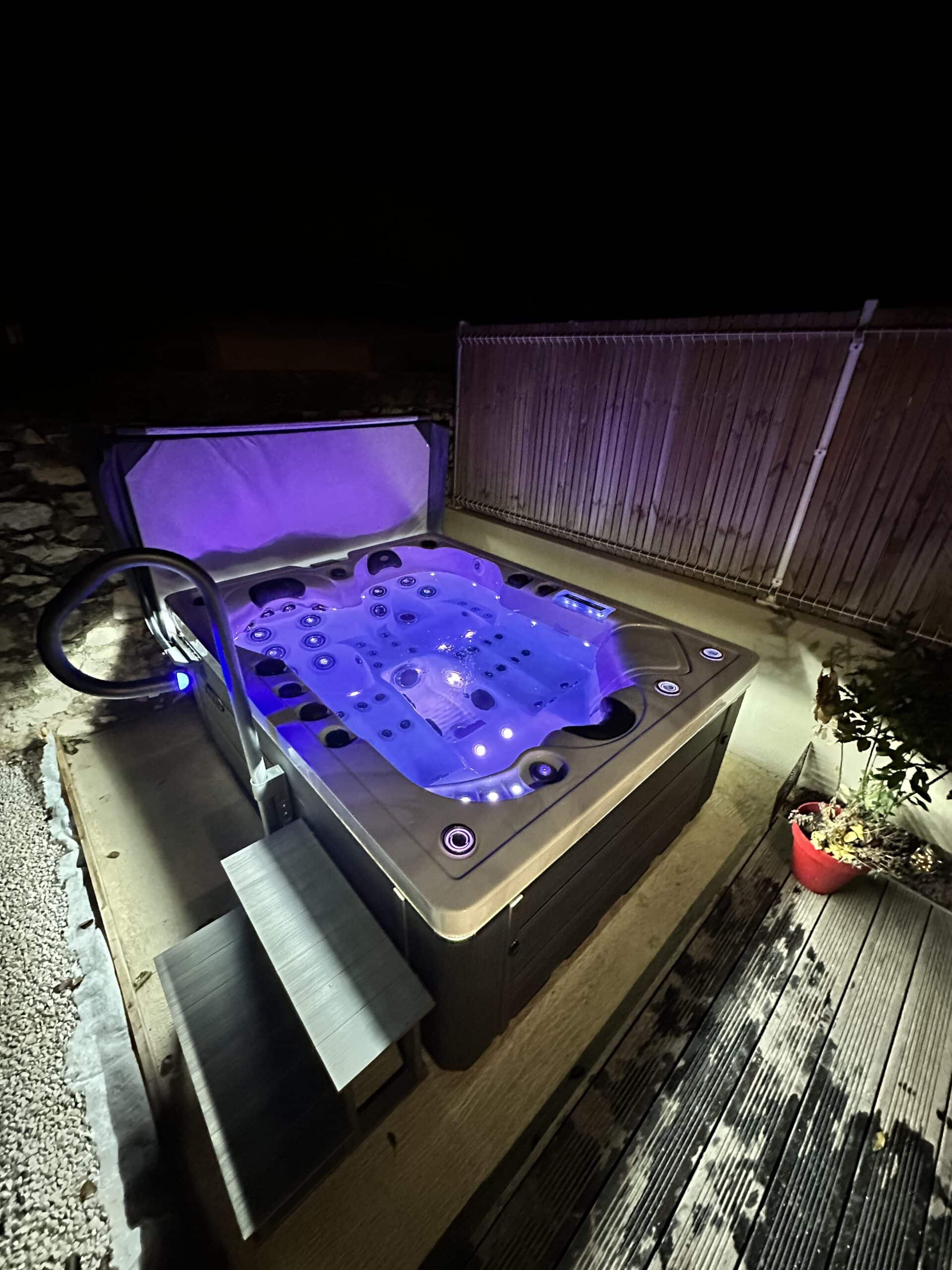 Spa extérieur moderne éclairé en bleu et violet la nuit. Installation de luxe sur une terrasse en bois avec marches et balustrade.