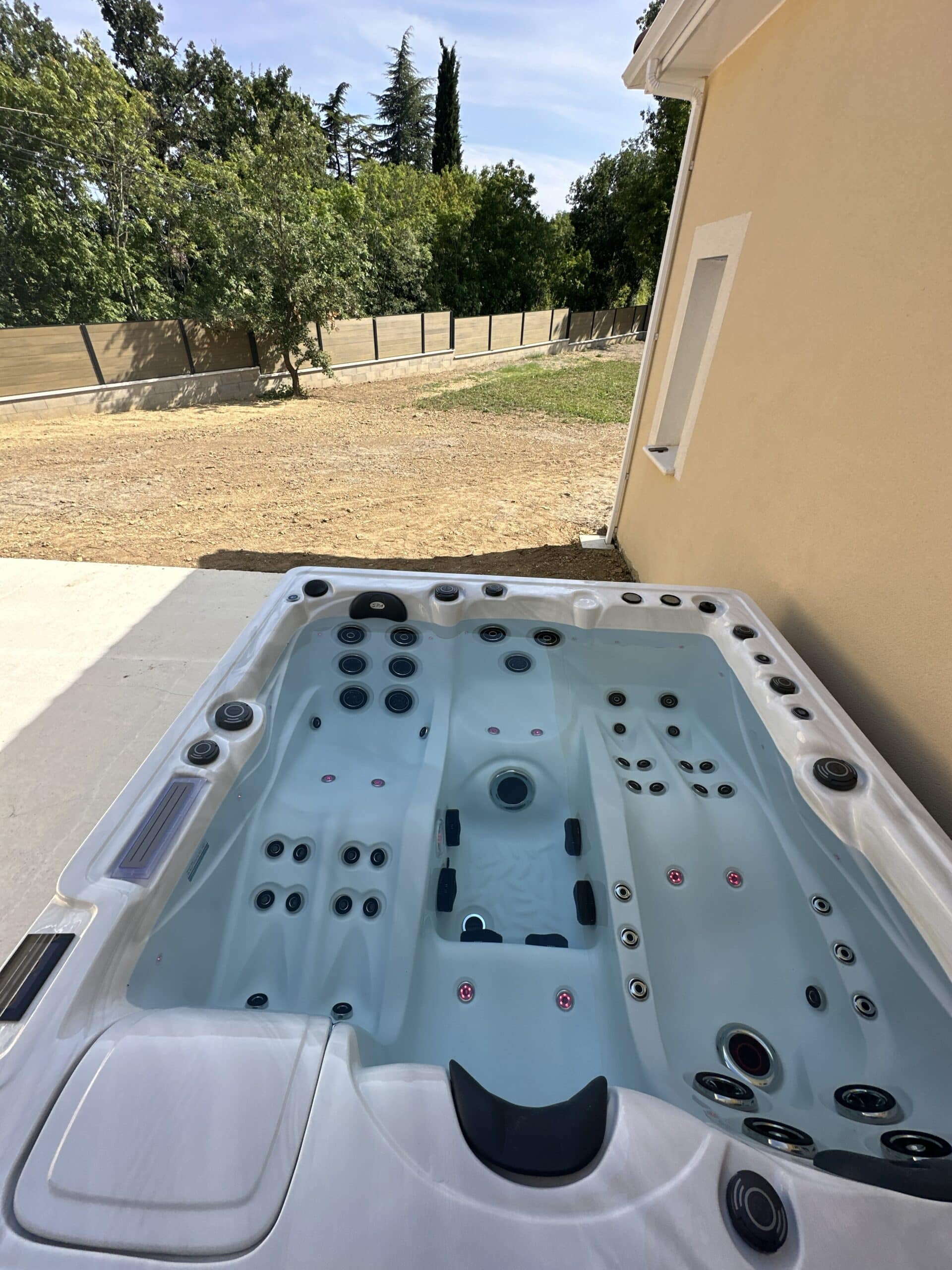 Spa extérieur blanc et jets noirs rempli d'eau claire, installé sur une terrasse face à un jardin en terre et des arbres.