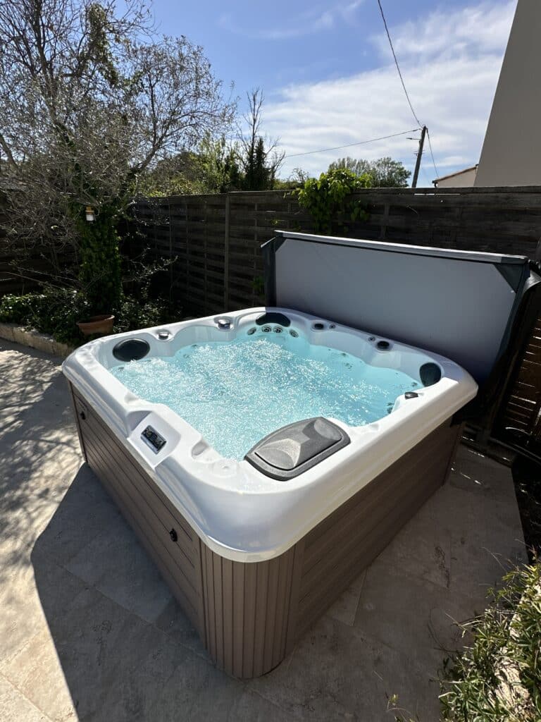 Grand spa extérieur blanc et marron avec jets en marche sur une terrasse ensoleillée. Clôture en bois et arbres à l'arrière.
