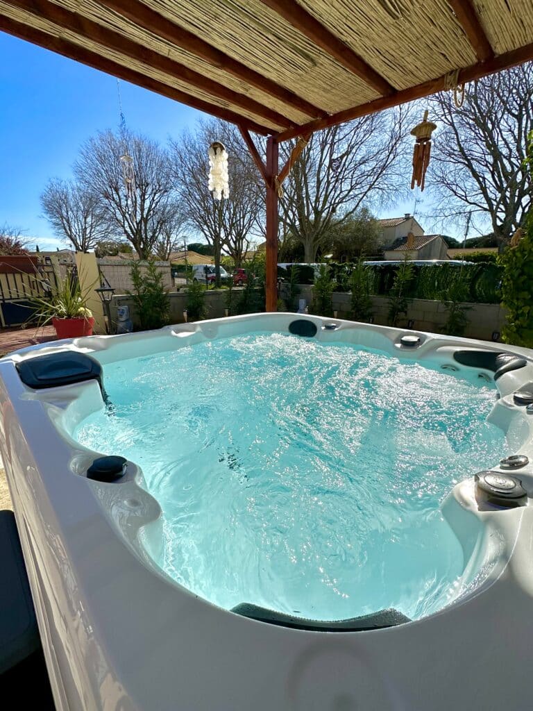 Grand spa extérieur blanc avec eau turquoise bouillonnante sous une pergola en bois et bambou par une journée ensoleillée.