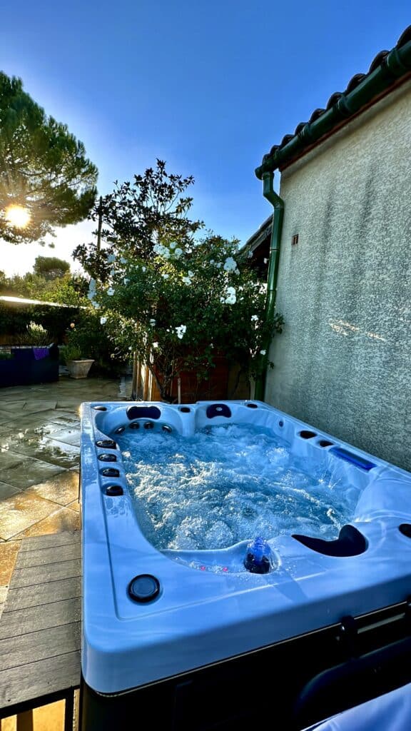 Spa moderne bouillonnant sur une terrasse extérieure au soleil couchant. Jardin luxuriant, mur de maison et roses blanches.