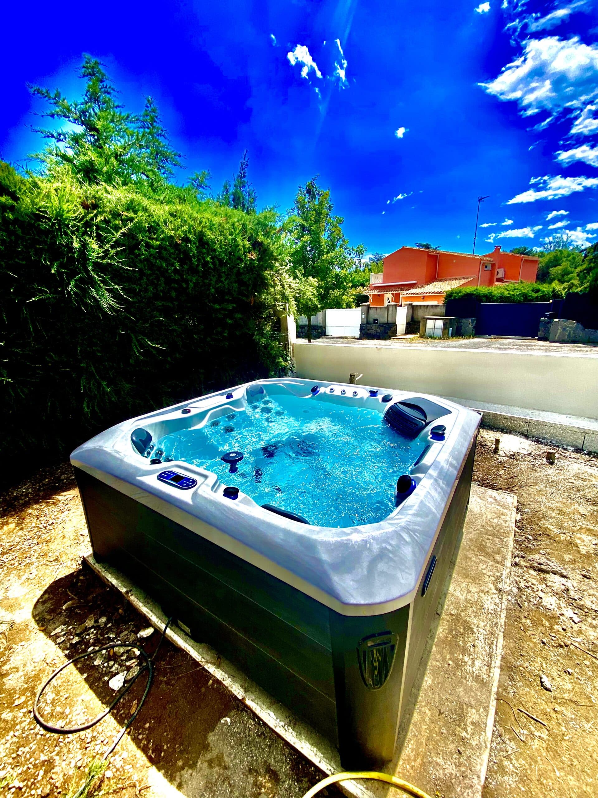 Spa extérieur en plein soleil avec eau bleue vive et bouillonnante, installé près d'une haie luxuriante et d'une maison orange sous un ciel azur.