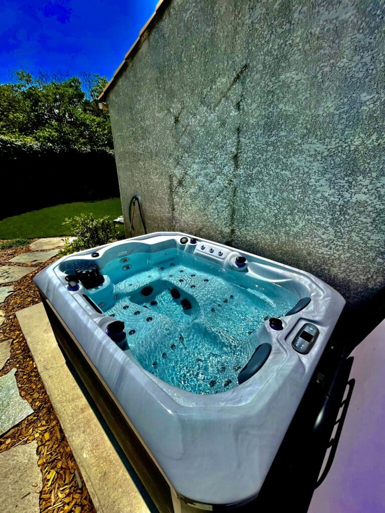 Jacuzzi spa extérieur moderne contre un mur texturé. L'eau turquoise pétille sous un ciel bleu vif. Détente au jardin.