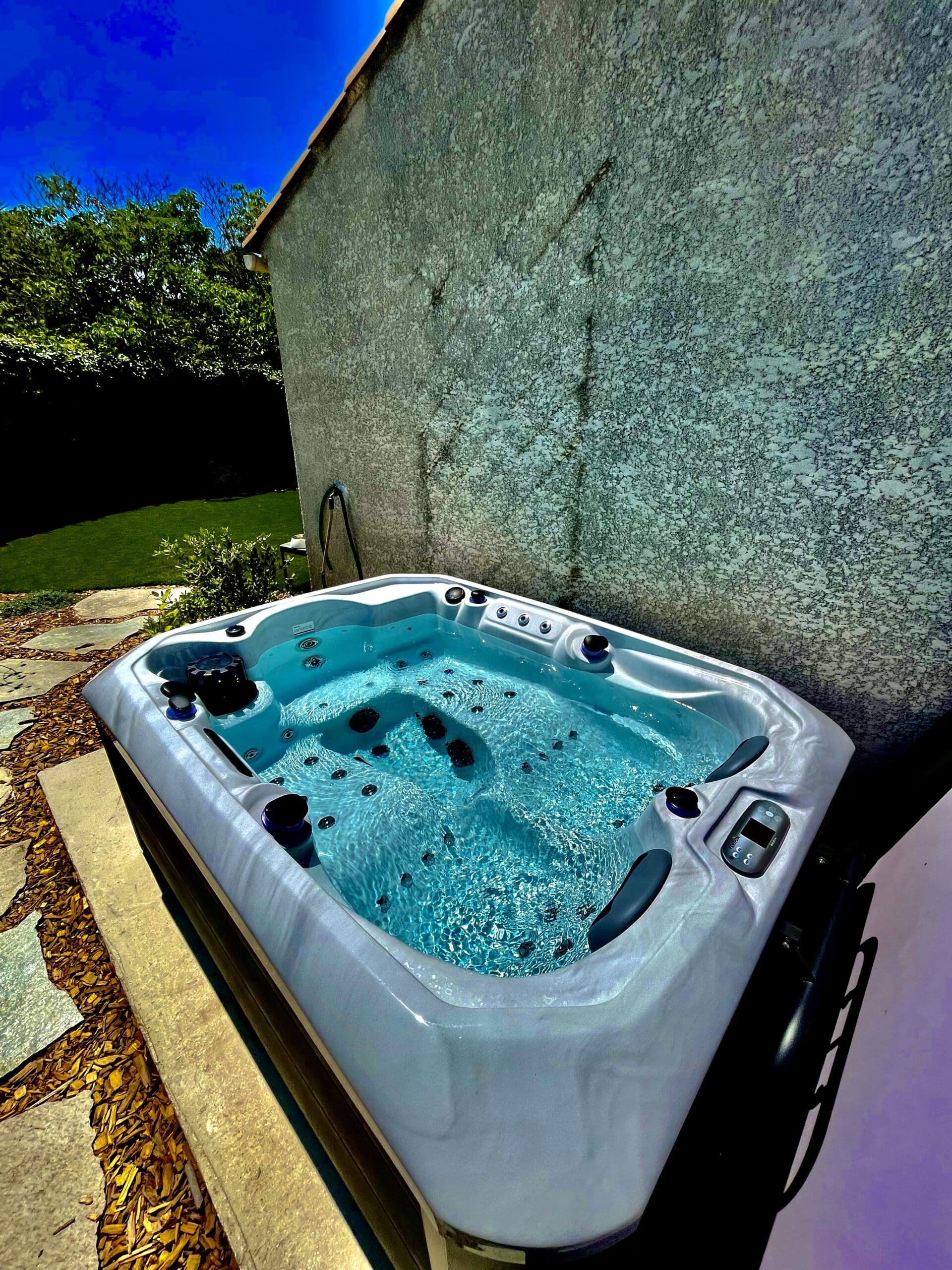 Jacuzzi spa extérieur moderne contre un mur texturé. L'eau turquoise pétille sous un ciel bleu vif. Détente au jardin.