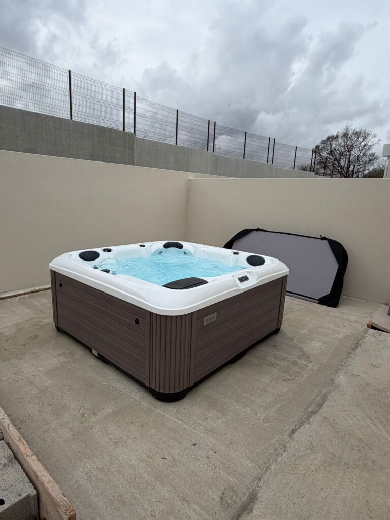 Installation de spa jacuzzi extérieur sur terrasse béton Spa jacuzzi extérieur marron et blanc, eau bouillonnante, installé sur une terrasse en béton. Murs hauts, clôture de sécurité et ciel gris.