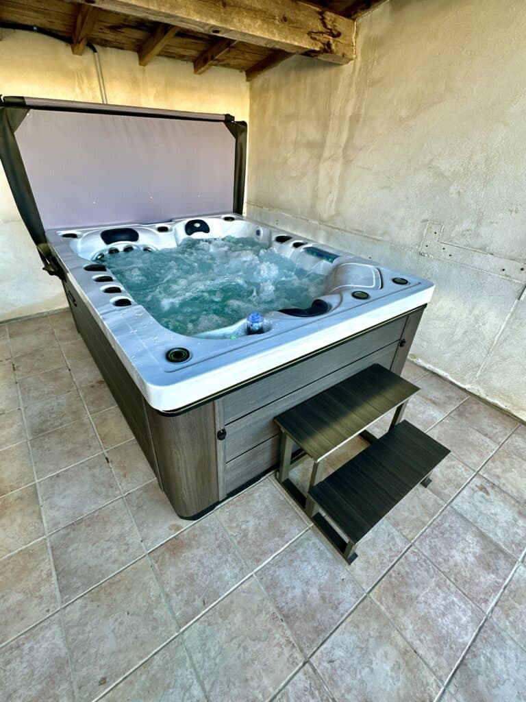 Spa extérieur moderne sous abri, prêt pour la détente Jacuzzi moderne gris foncé avec jets actifs, installé sous un plafond de poutres en bois. Marches noires sur carrelage clair.