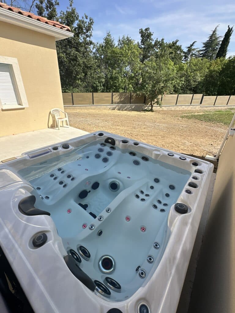 Spa extérieur neuf : détente et jets sur la terrasse. Spa extérieur haut de gamme neuf avec multiples jets et éclairage LED, installé sur une terrasse près d'une maison et d'un jardin.