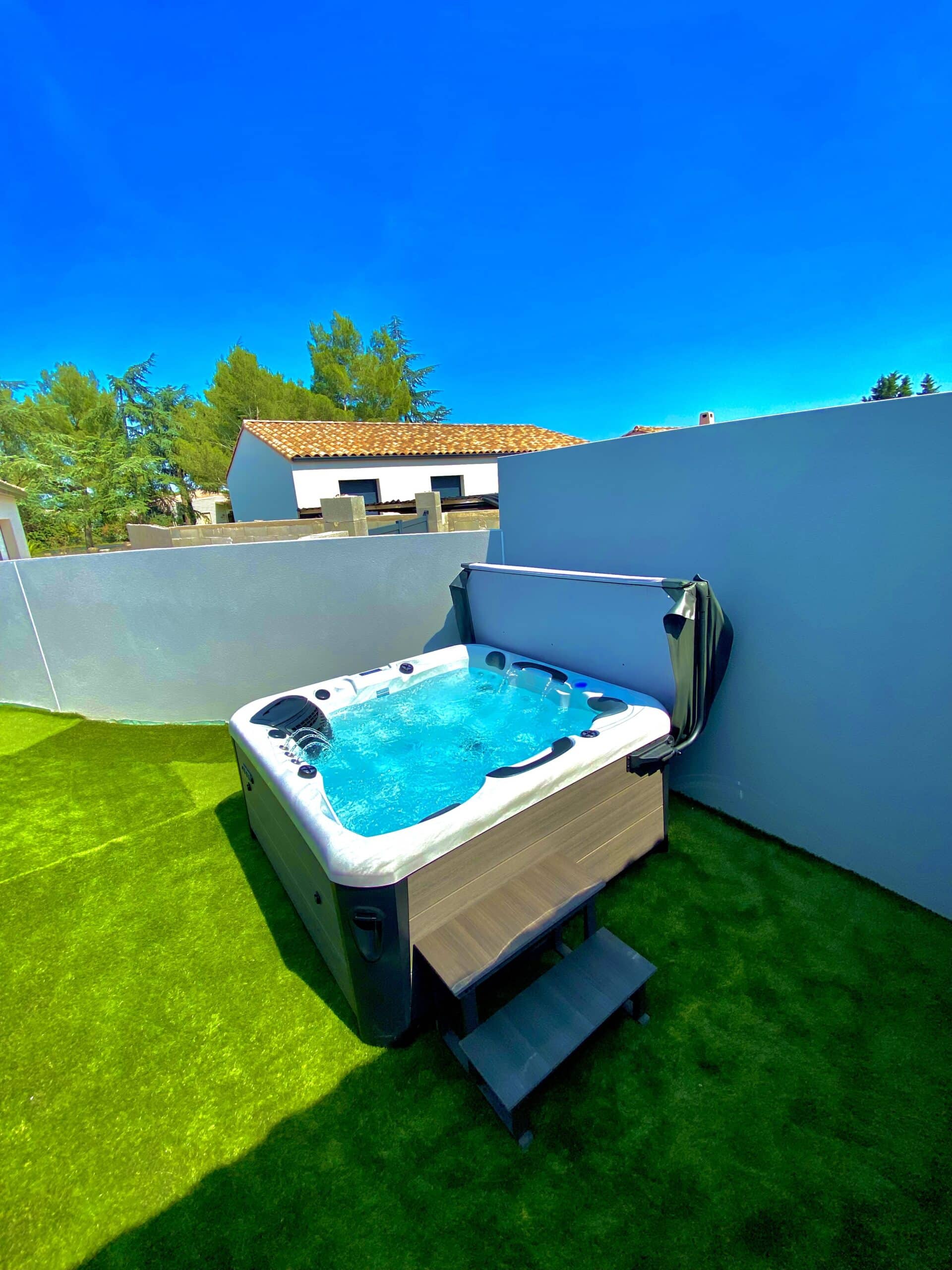 Spa de luxe bouillonnant sur terrasse en gazon synthétique. Ciel bleu éclatant, mur d'intimité gris-bleu et maison méditerranéenne.