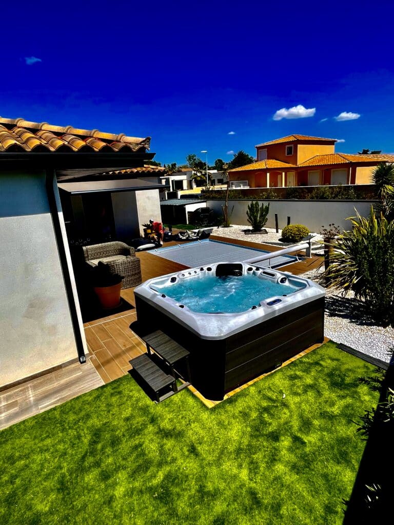 Spa extérieur luxe : Détente garantie sous le ciel bleu Luxueux spa extérieur bouillonnant sur une terrasse en bois. Jardin en gazon vert, murs blancs, toits terracotta sous ciel bleu.