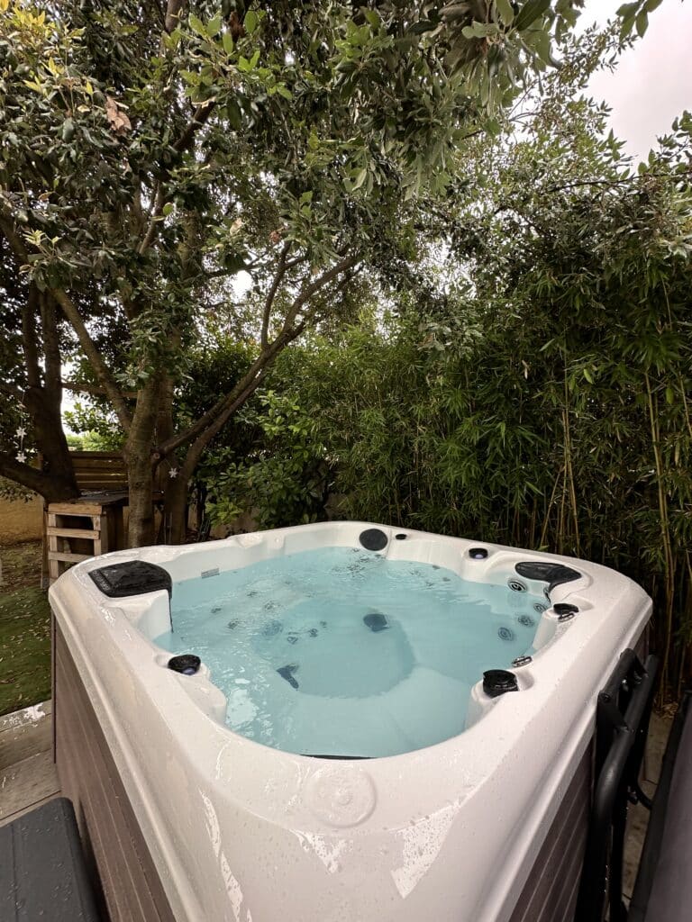 Spa extérieur dans jardin ombragé | Détente & ambiance bambou. Spa extérieur blanc entouré d'une végétation dense : grands arbres à gauche et haie de bambous à droite. L'eau turquoise invite à la détente.