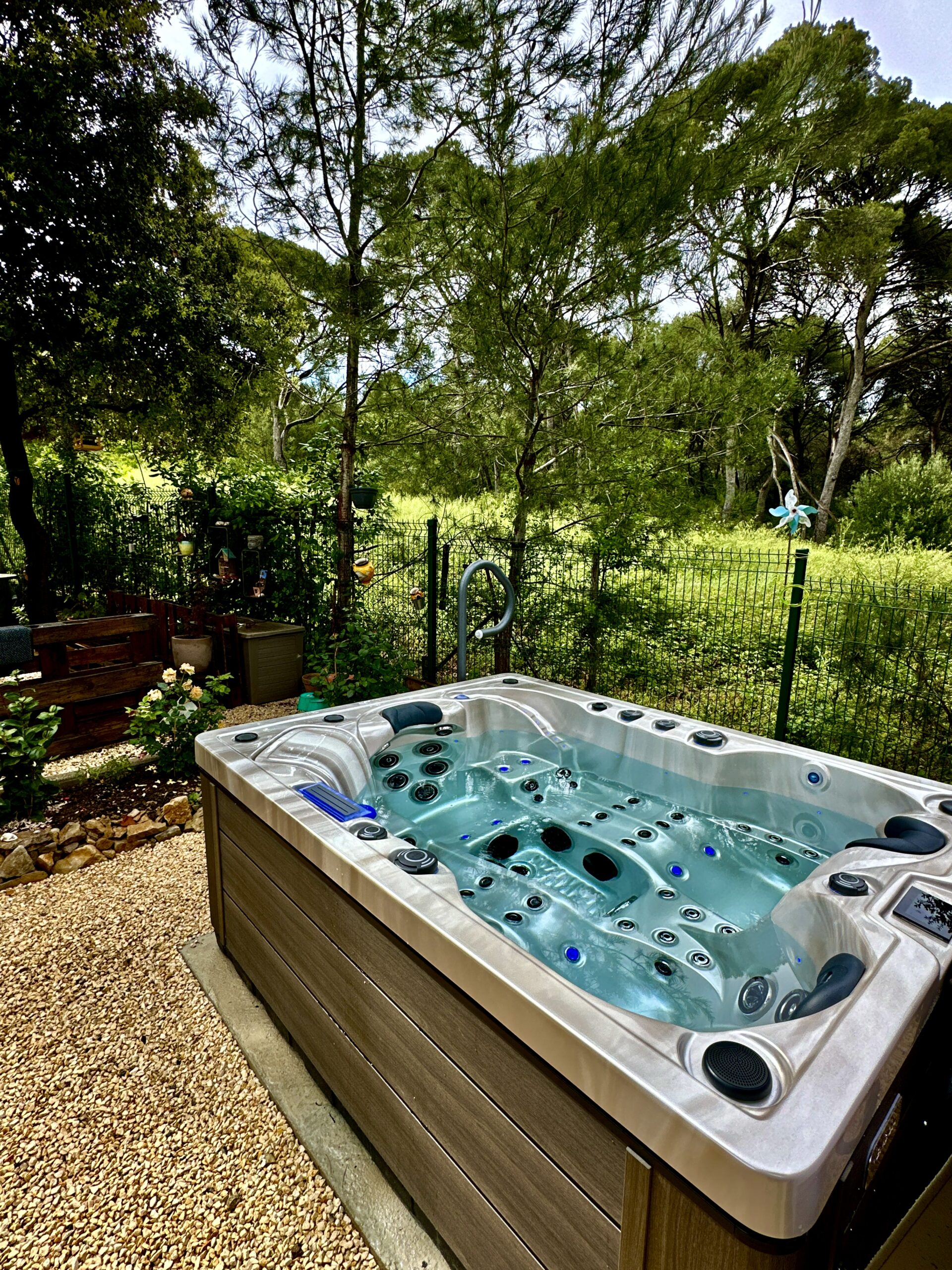 Grand spa extérieur sur gravier, avec boîtier en bois composite brun, eau turquoise. Jardin luxuriant et arbres en arrière-plan.