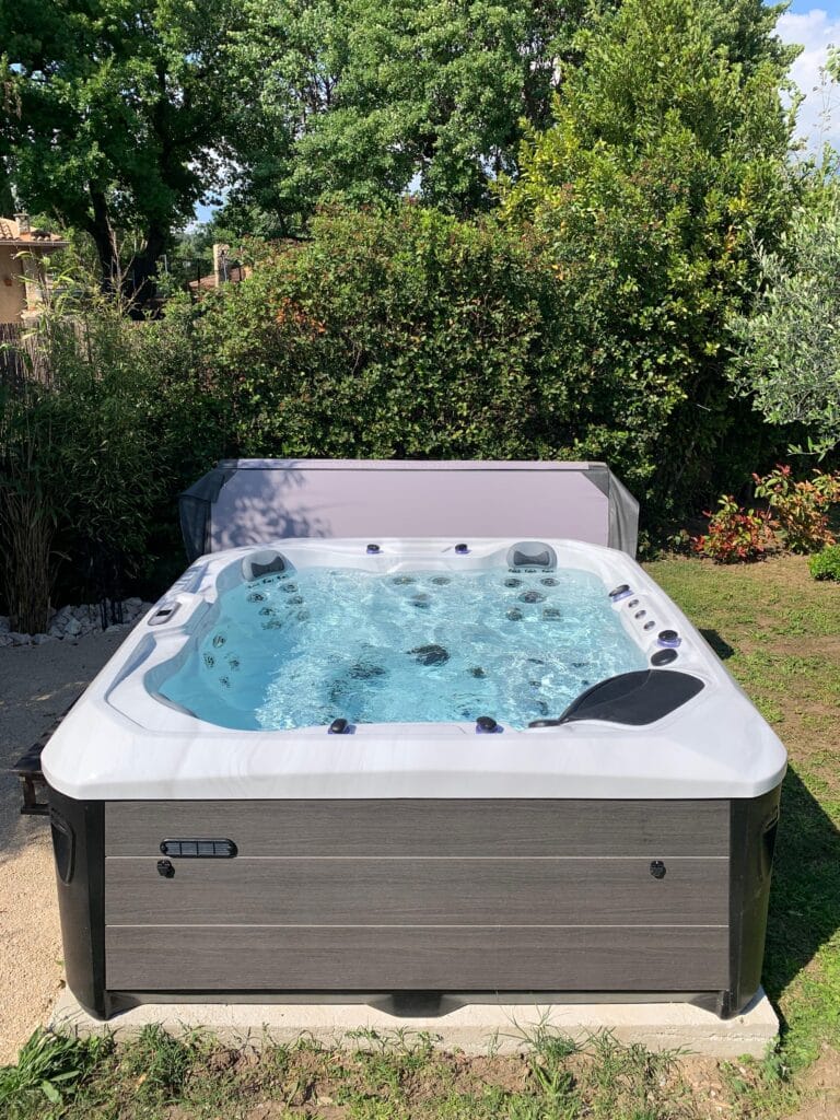 Spa carré moderne en bois composite gris foncé avec eau bleue bouillonnante. Installation extérieure dans un jardin privé luxuriant.