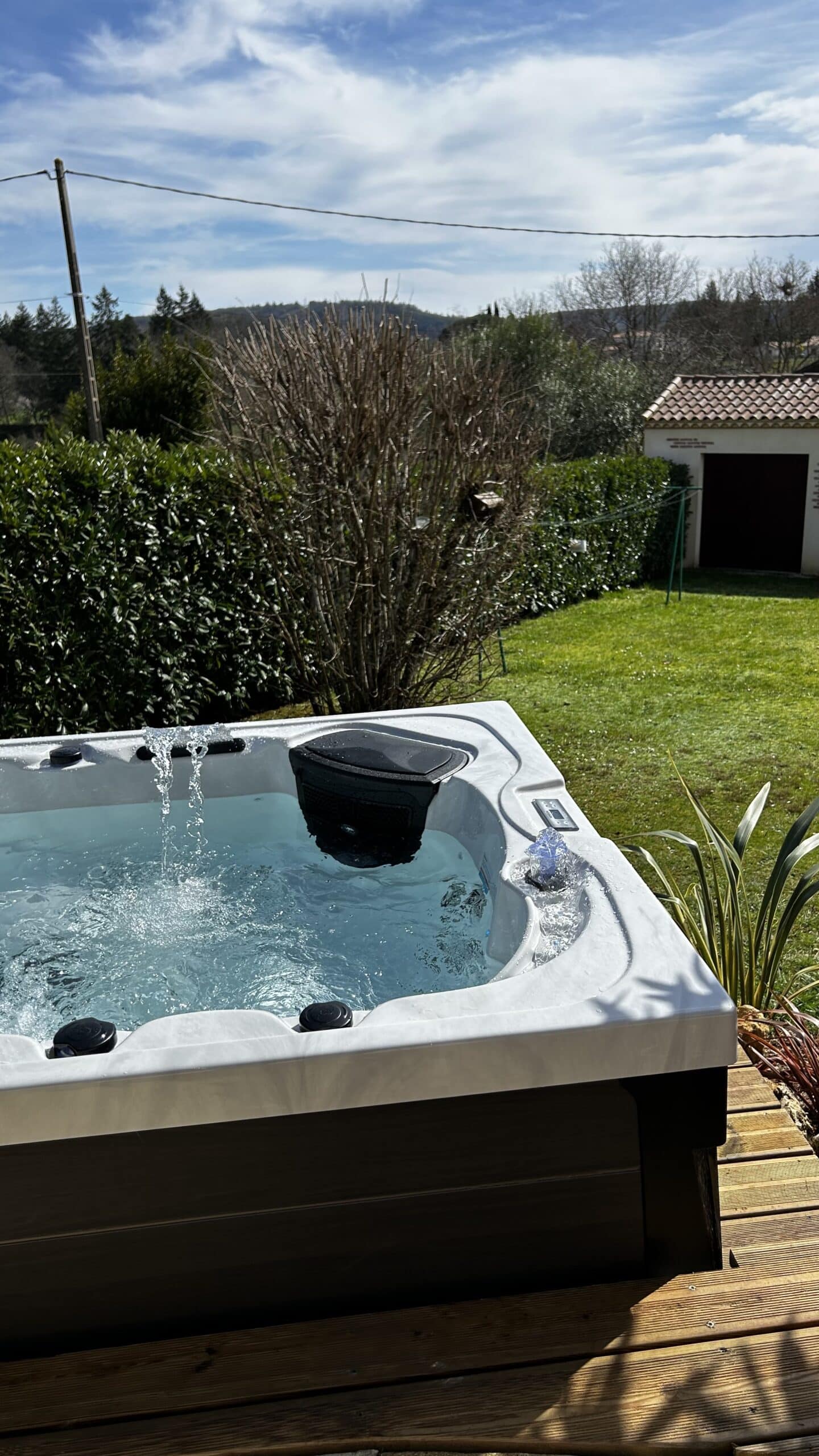 Bain à remous extérieur blanc avec cascade d'eau dans un jardin. Terrasse en bois, haie verte et ciel bleu nuageux.
