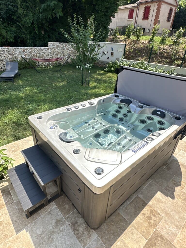 Spa extérieur moderne gris avec marches en composite, posé sur une terrasse carrelée ensoleillée, avec jardin, hamac et maison en arrière-plan.