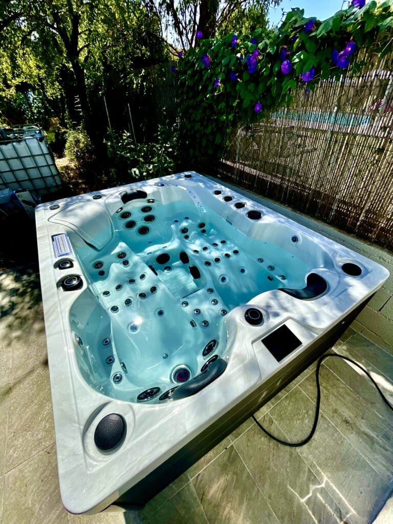 Jacuzzi extérieur moderne et lumineux sur terrasse carrelée, bordé de haies vertes et de fleurs violettes sous un soleil éclatant.