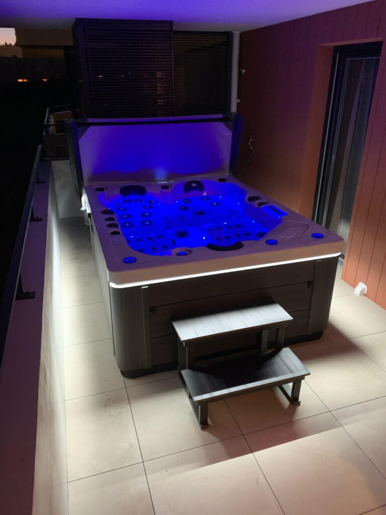 Spa de luxe sur balcon de nuit, baignoire éclairée par LED bleu intense. Bain à remous moderne avec habillage gris et marchepied.