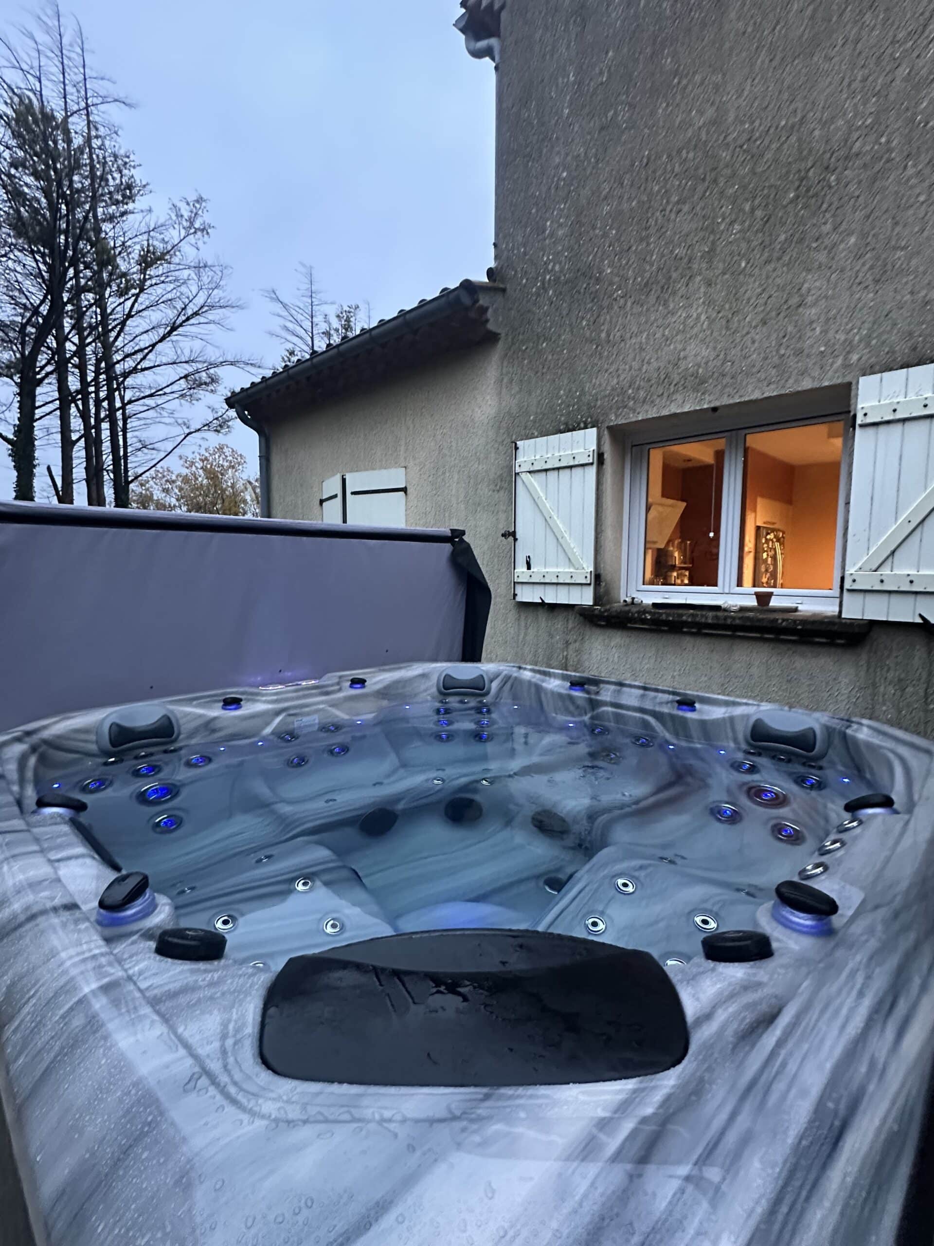 Luxueux spa d'extérieur au crépuscule. L'eau bleue éclairée contraste avec le mur de stuc et la cuisine intérieure chaleureuse.