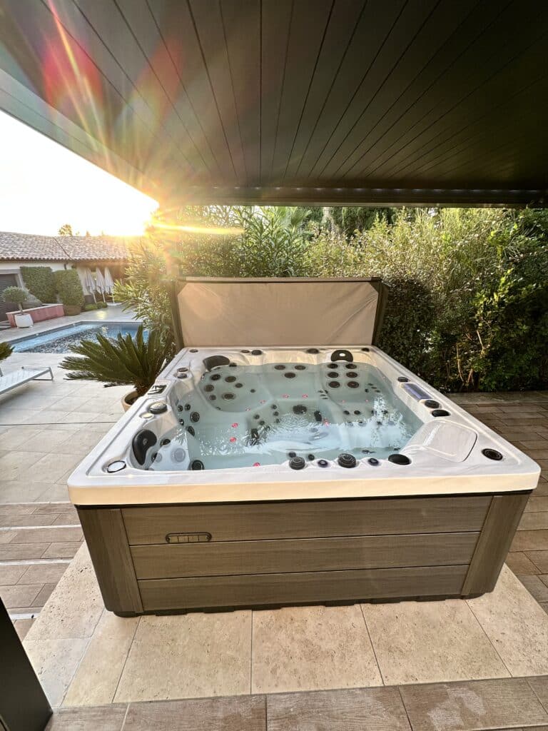 Spa de luxe extérieur, couleur bois, sous une pergola sombre au coucher du soleil. Forte lumière et reflets arc-en-ciel sur le plafond. Piscine en arrière-plan.