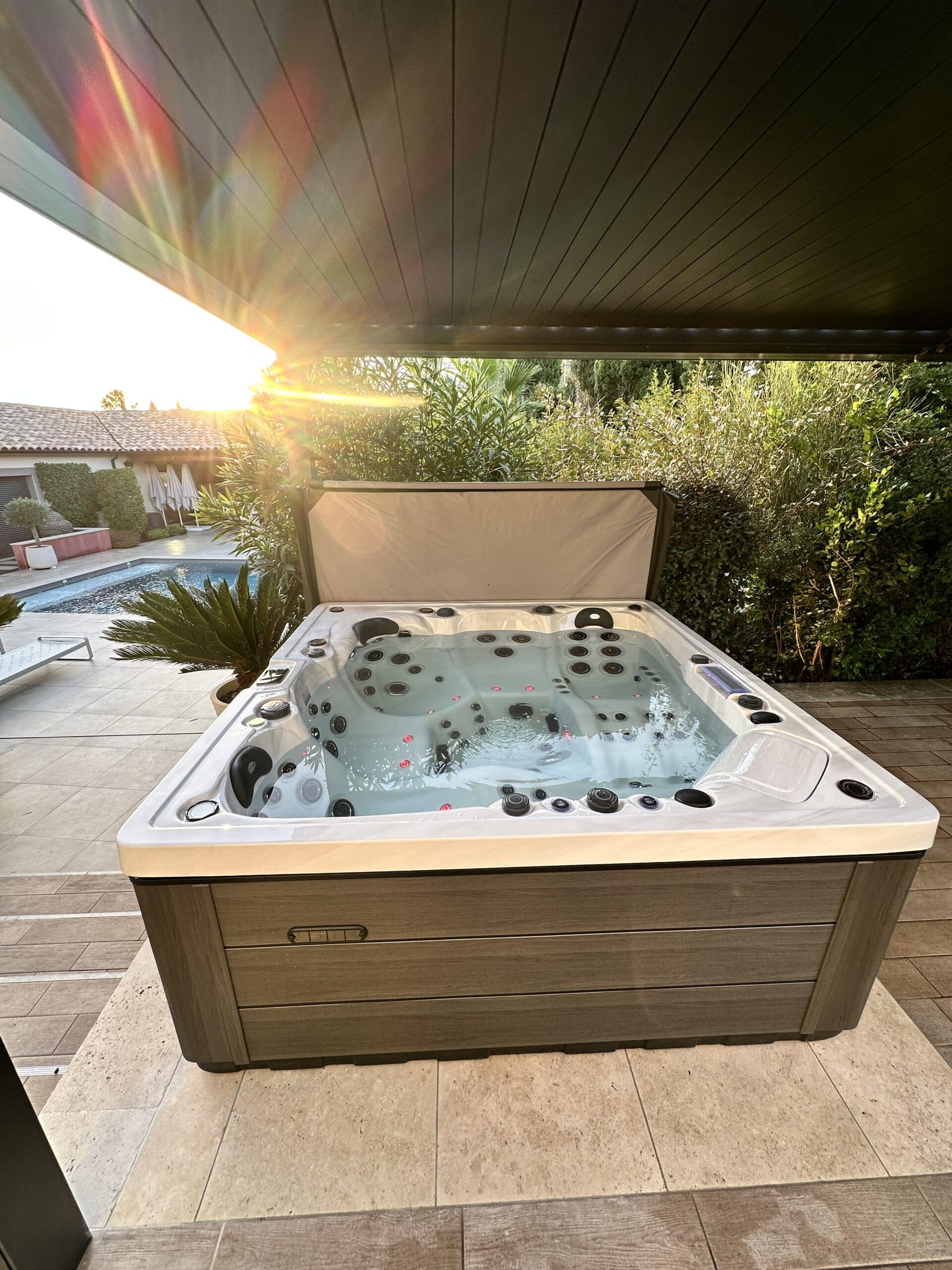 Spa de luxe extérieur, couleur bois, sous une pergola sombre au coucher du soleil. Forte lumière et reflets arc-en-ciel sur le plafond. Piscine en arrière-plan.