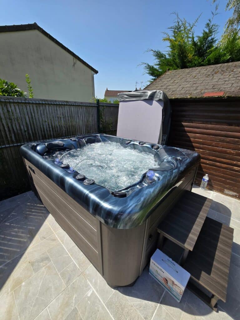 Spa extérieur Vendom en bulles : votre jacuzzi de jardin idéal Jacuzzi spa Vendom moderne extérieur en marche, coque bleu marbré. Bulles, escalier et couvercle relevé. Ciel bleu éclatant.