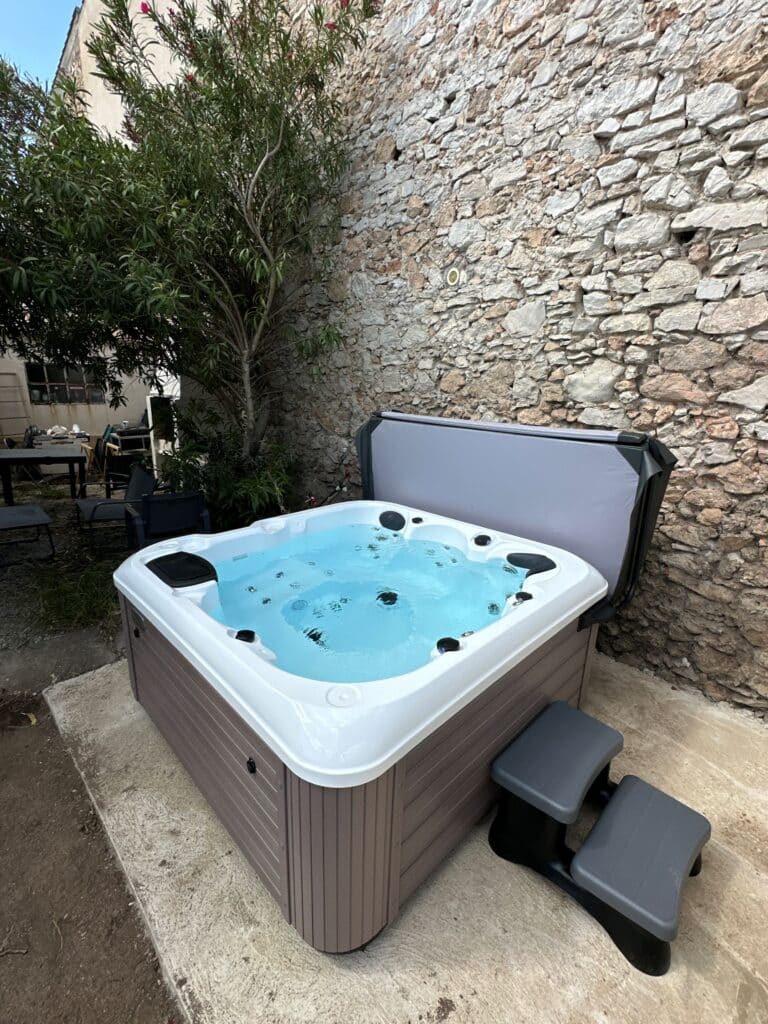 Spa jacuzzi extérieur pour détente dans un cadre rustique Spa jacuzzi moderne, jupe marron, avec eau bouillonnante installé dehors. Il est adossé à un grand mur de pierre rustique près d'un arbre.