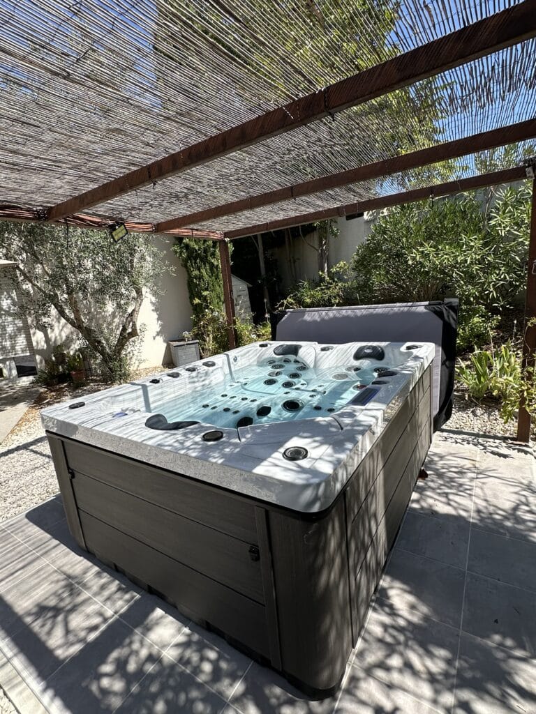 Spa Jacuzzi extérieur sous pergola bambou | Détente jardin Jacuzzi moderne extérieur sous une pergola en bambou ombragée. La cuve blanche et le revêtement foncé sont entourés d'un jardin méditerranéen.