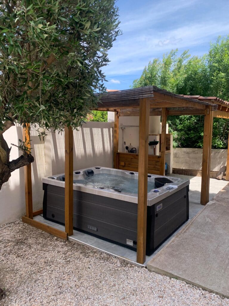 Spa extérieur sous pergola bois : Profitez de la détente. Spa de jardin moderne sous une pergola en bois rustique. L'olivier et les murs blancs créent une ambiance méditerranéenne ensoleillée.