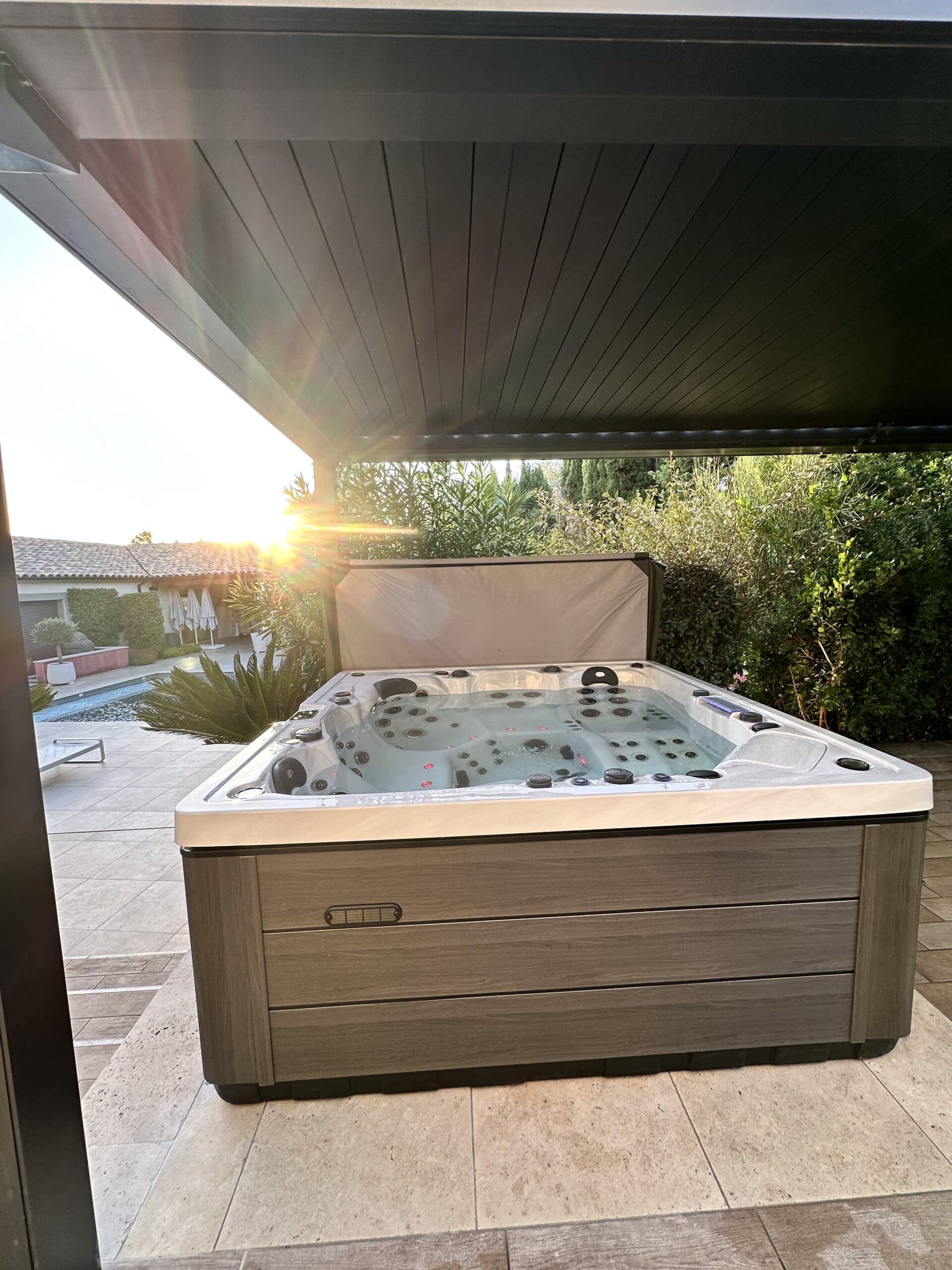 Spa de luxe moderne sur une terrasse pavée, sous une pergola contemporaine, avec vue sur la piscine au coucher du soleil et reflets lumineux.