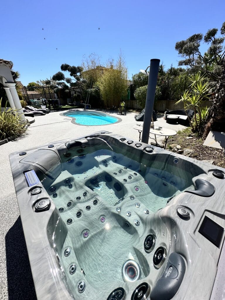 Spa extérieur moderne gris marbré rempli d'eau, installé sur une terrasse en gravier près d'une petite piscine turquoise sous un ciel bleu d'été.
