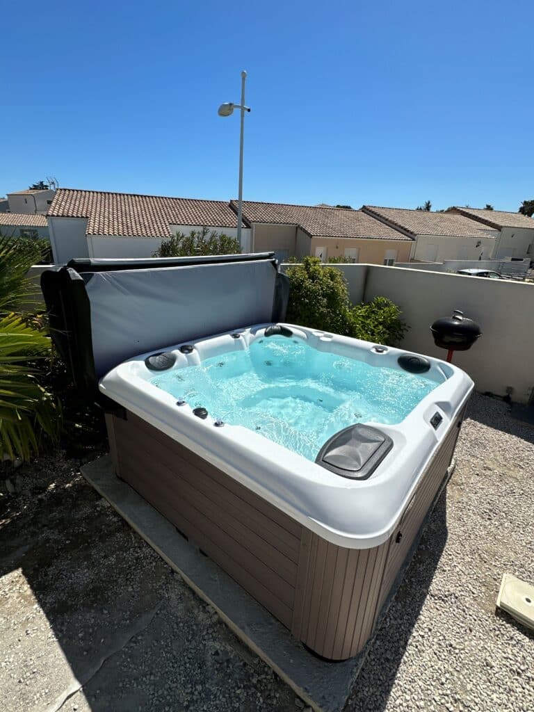 Spa Jacuzzi extérieur : Profitez de la détente sous le soleil Grand spa jacuzzi extérieur en plein soleil, avec eau turquoise bouillonnante. La cuve est posée sur une terrasse résidentielle en gravier.