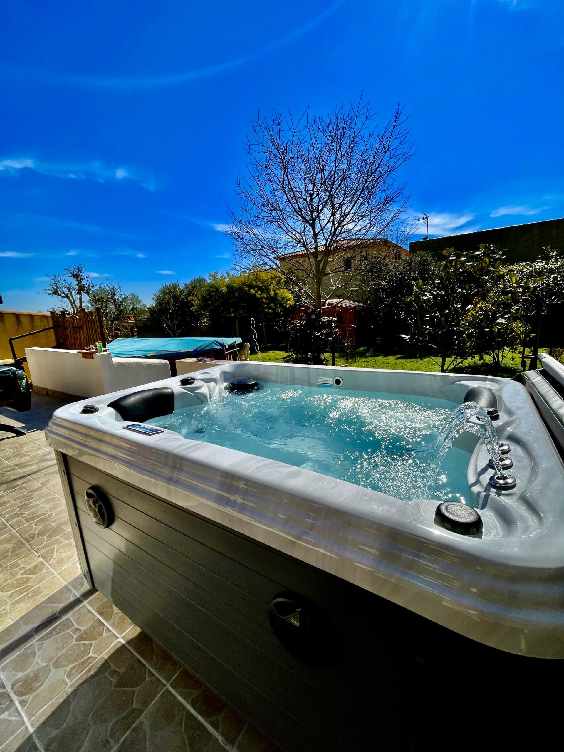 Spa extérieur moderne avec jets d'eau pétillante sur une terrasse carrelée. Ciel bleu éclatant et végétation en arrière-plan.