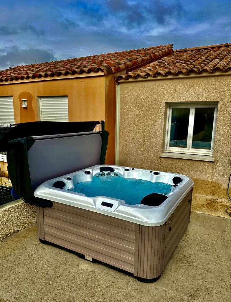 Jacuzzi extérieur : détente sur terrasse sous ciel nuageux. Spa extérieur moderne bouillonnant, couverture ouverte, sur terrasse bétonnée. Murs ocre, toit en tuiles terre cuite sous un ciel bleu dramatique.