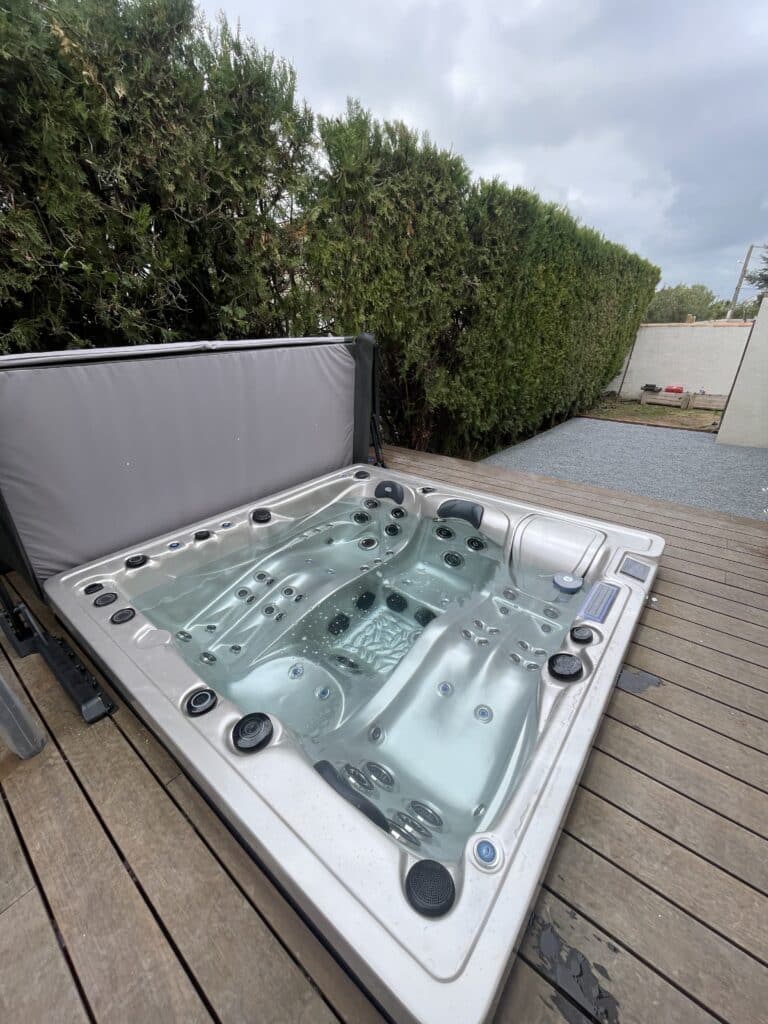 Jacuzzi moderne intégré dans une terrasse en bois, couverture ouverte. Bain à remous prêt à l'emploi avec haie extérieure.