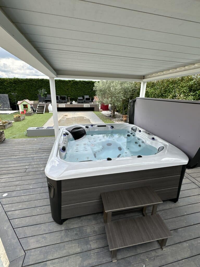 Spa jacuzzi moderne sous pergola sur une terrasse bois sombre. Le jardin aménagé avec haie, olivier et aire de jeu est visible à l'arrière.