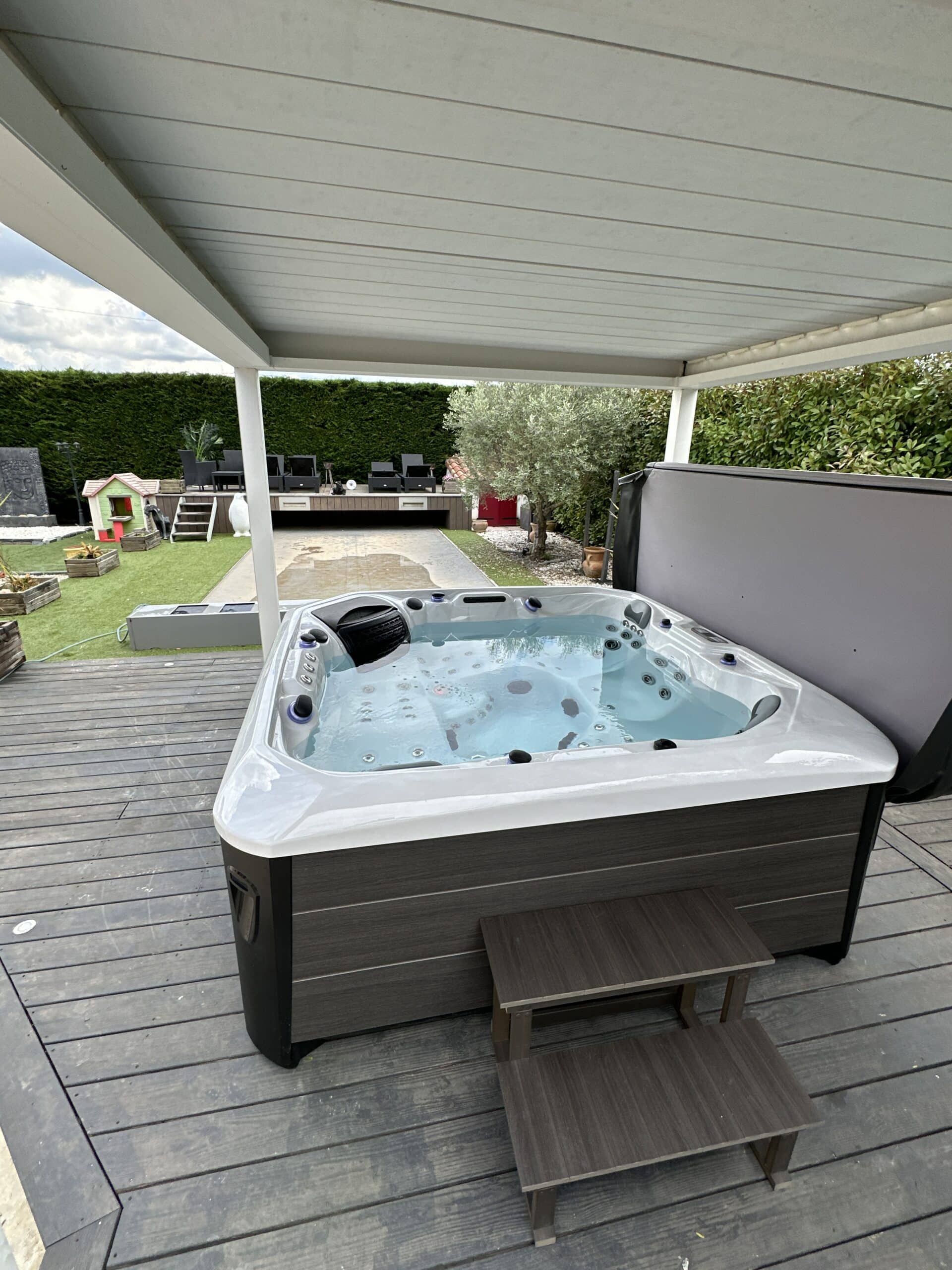 Spa jacuzzi moderne sous pergola sur une terrasse bois sombre. Le jardin aménagé avec haie, olivier et aire de jeu est visible à l'arrière.