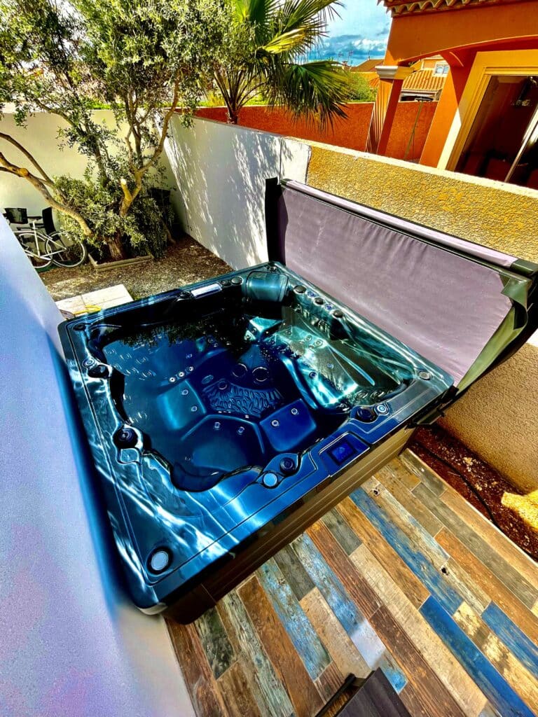 Jacuzzi extérieur bleu sur une terrasse ensoleillée aux murs clairs et oranges. Sol en lattes de bois rustiques et végétation.