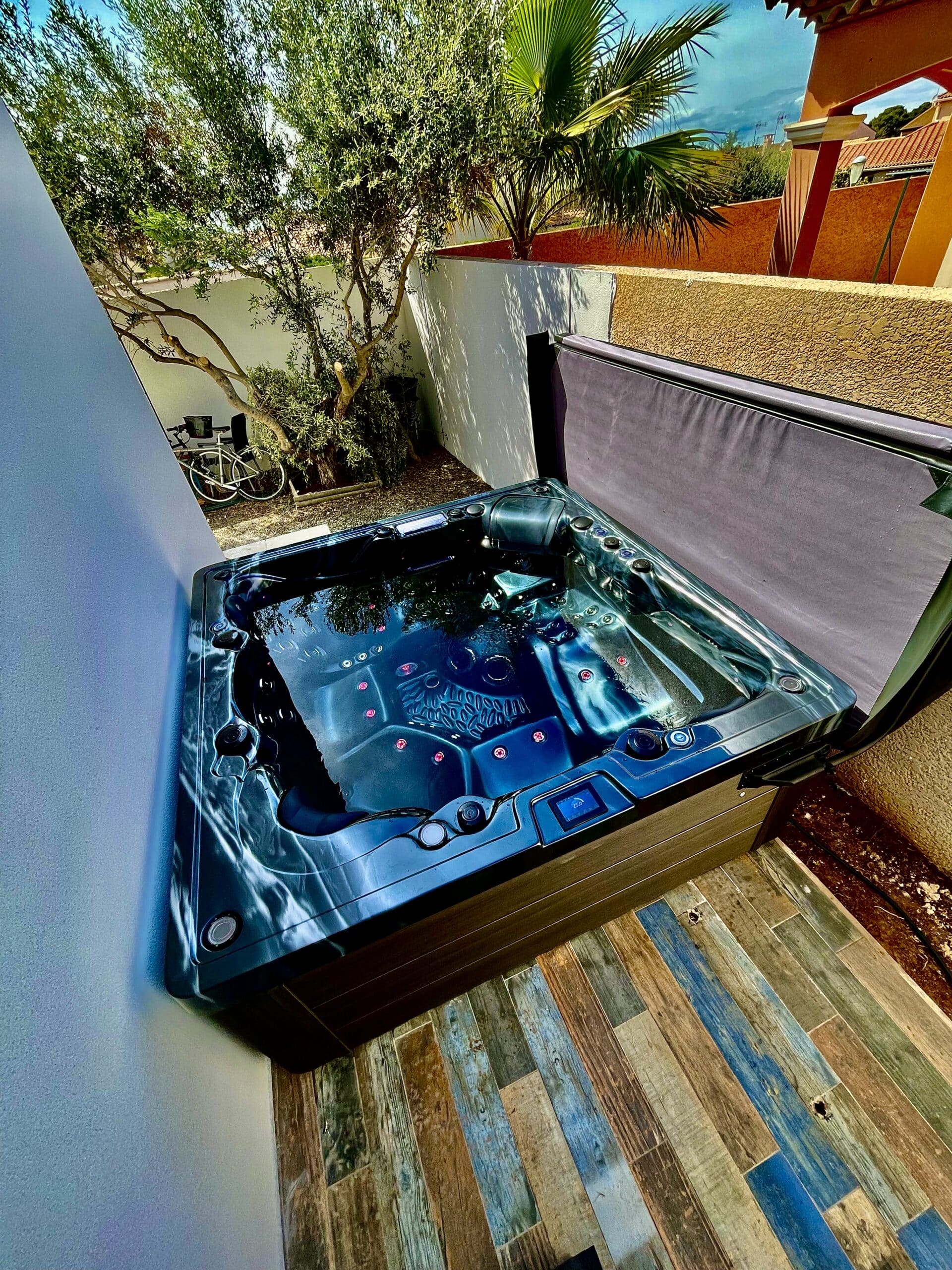 Spa jacuzzi moderne bleu extérieur ouvert sur une terrasse en bois patiné bleu et brun. Végétation luxuriante, contrôle affichant 25.0°C.