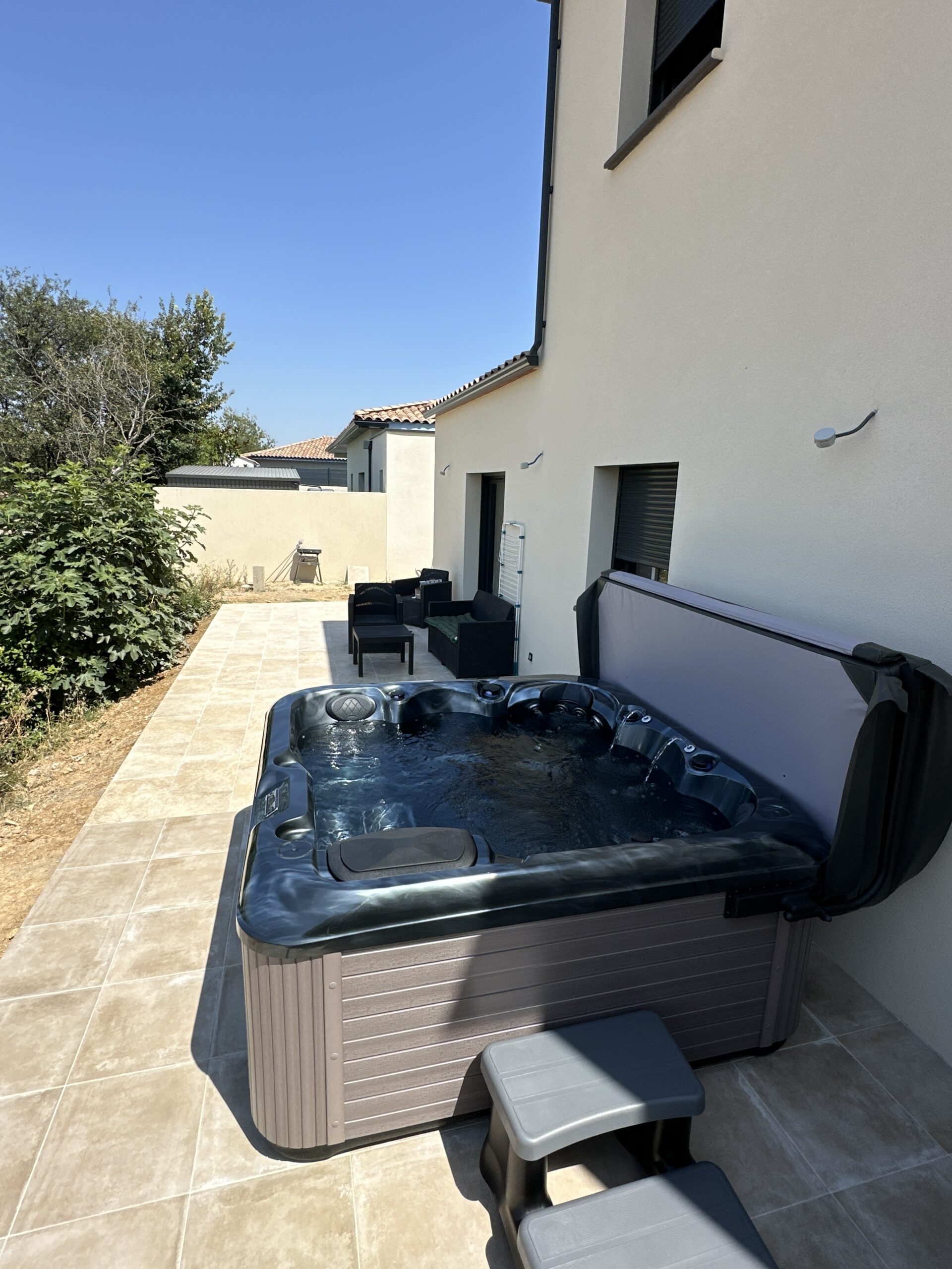 Grand spa jacuzzi extérieur foncé sur terrasse carrelée beige. Murs de maison modernes et ciel bleu en plein soleil d'été.