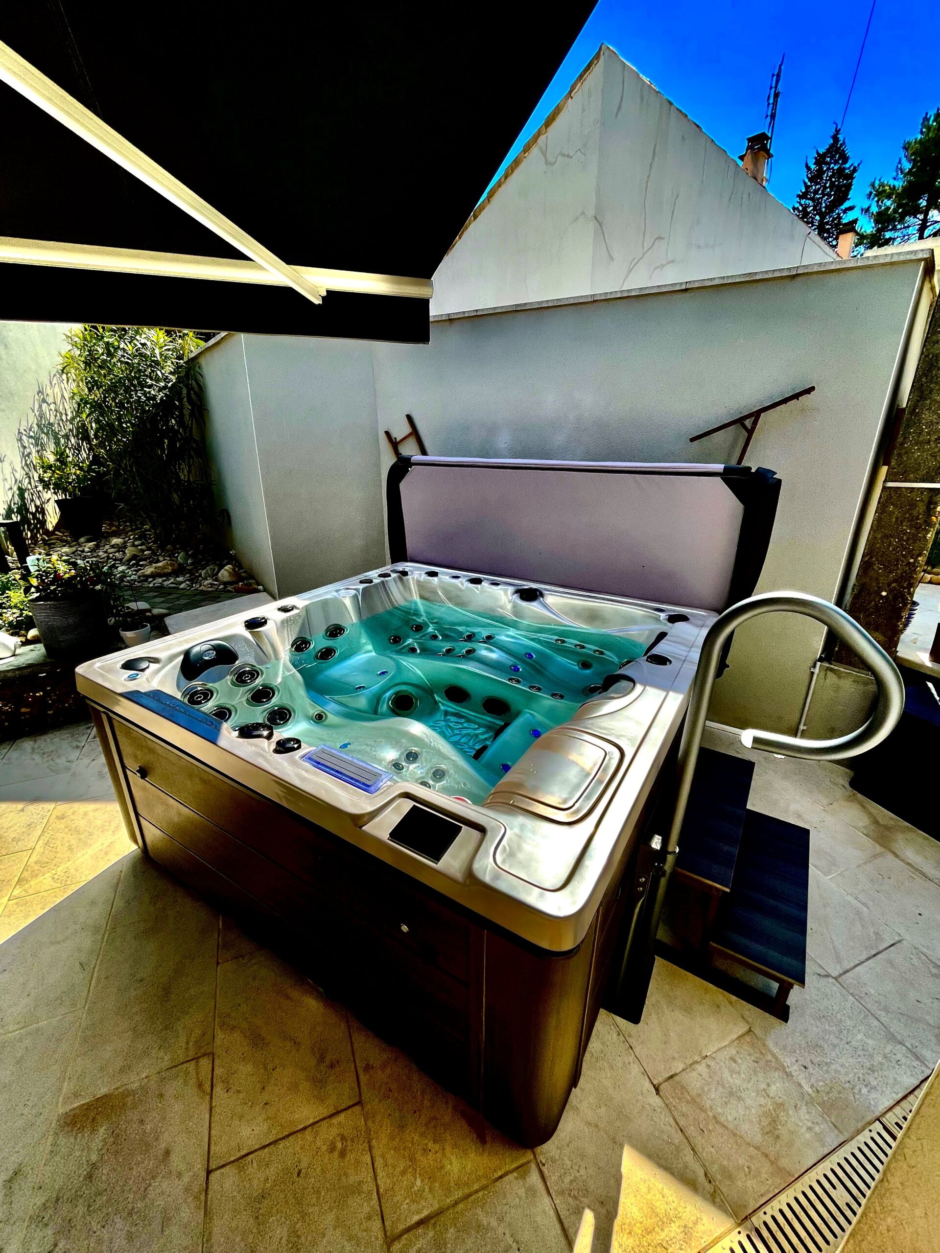 Luxueux spa extérieur sur terrasse carrelée, sous auvent, avec eau turquoise bouillonnante. Couvercle levé et rampe d'accès visible.