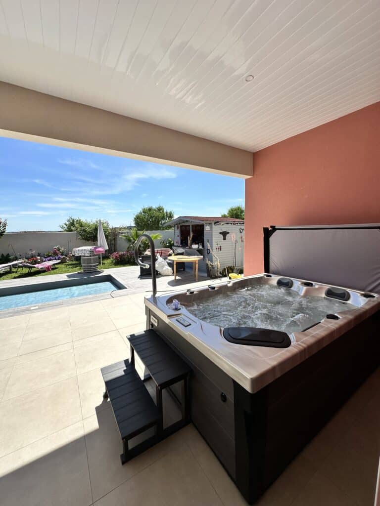 Jacuzzi extérieur sur terrasse couverte avec vue sur la piscine Spa jacuzzi extérieur bouillonnant installé sur une terrasse couverte. Vue sur la piscine, le jardin et le ciel bleu.
