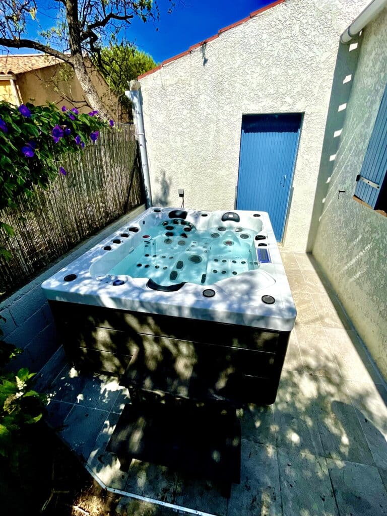 Spa extérieur sur terrasse ensoleillée et ambiance détente Jacuzzi extérieur moderne sur une terrasse ensoleillée. Mur blanc, porte bleue vibrante et fleurs pourpres. Espace privatif de détente.
